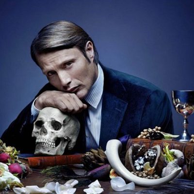 Mads Mikkelsen as Hannibal Lecter rests his head on a skull as he sits at a table adorned with flowers and wine