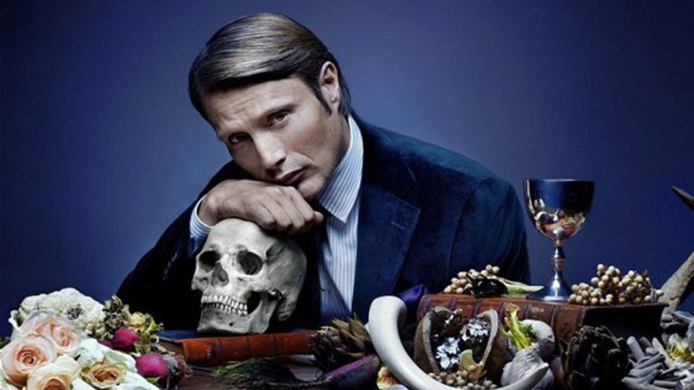 Mads Mikkelsen as Hannibal Lecter rests his head on a skull as he sits at a table adorned with flowers and wine