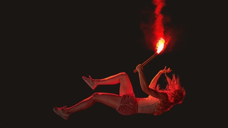 A girl falls against a black background, with a lit flare in her hand
