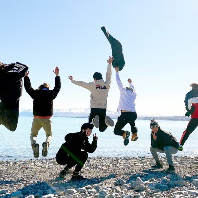 Five BTS members jump into the air while two crouch down for a picture in front of a New Zealand lake in BTS Bon Voyage Season 4