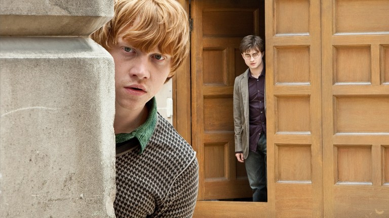 Ron Weasley peeks out from around the corner, as Harry Potter stands in a doorway in the background