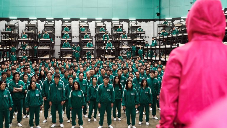 Squid Game competitors stand in a crowd as a man in a red jumpsuit looks on