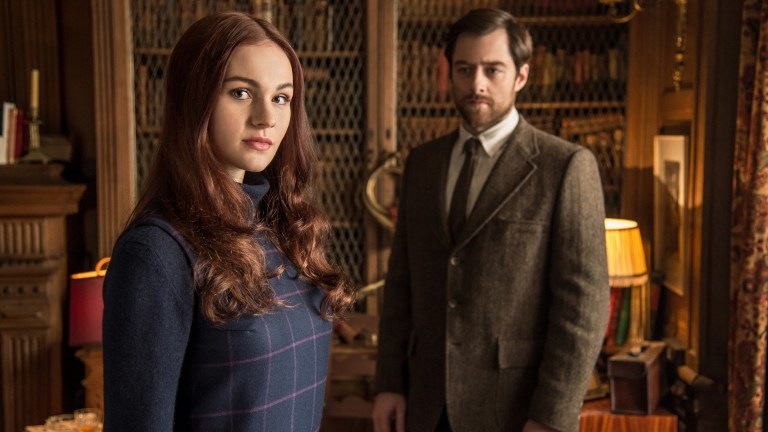Brianna stands in the foreground of a library as Roger looks at her in Outlander