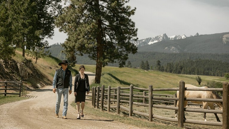 John Dutton (Kevin Costner) and a woman walk down a dirt road in beautiful Montana scenery on Yellowstone.