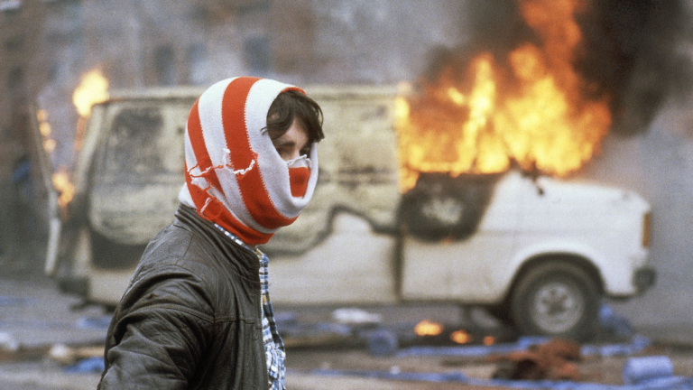 A youth in front of a blazing vehicle in Belfast during The Troubles