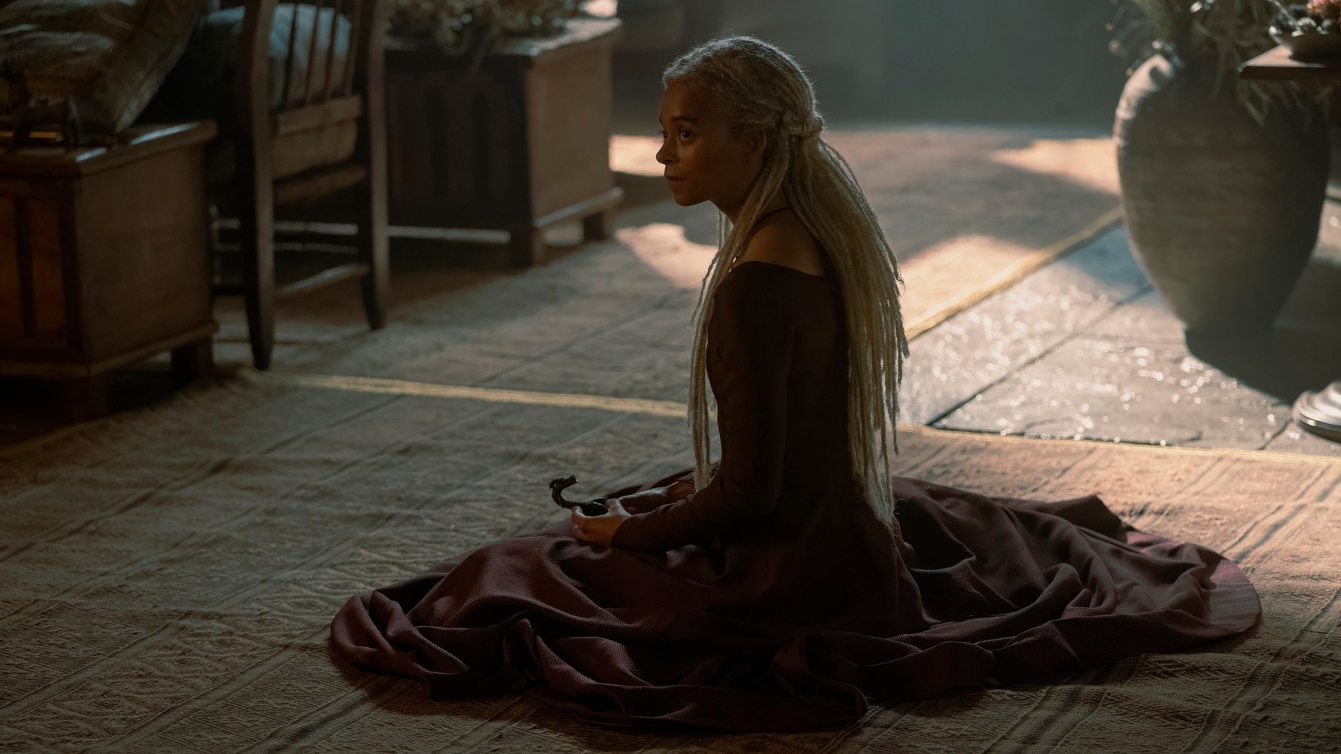 A woman (Rhaena) with long blonde dreads sits on a rug, with her maroon dress pooled around her. She is looking to the left off-screen. There is a potted plant behind her to the right and a chair and two tables to her left.