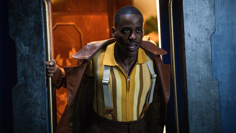 Ncuti Gatwa as the Doctor in a leather jacket, tweed trousers and braces looking out of a train door