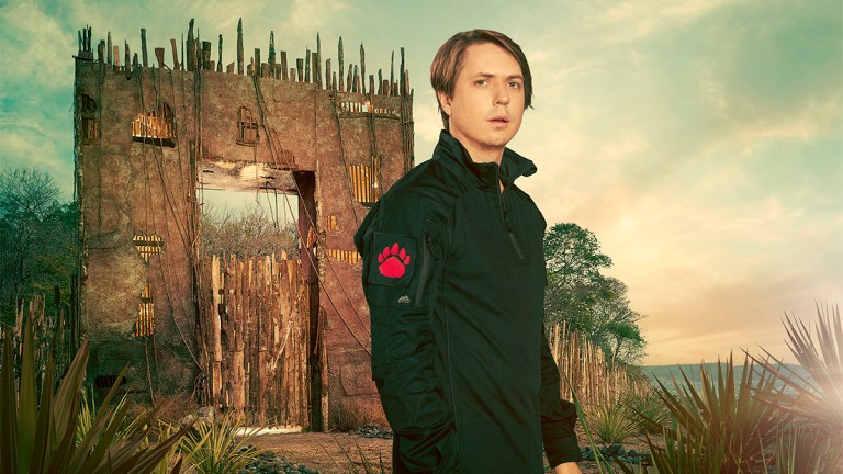 Joe Thomas wearing a dark green jumpsuit and standing next to a large wooden gate