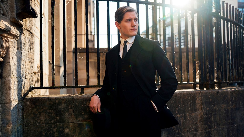 Adam Hugill in a black 1920s-style suit standing against some railings