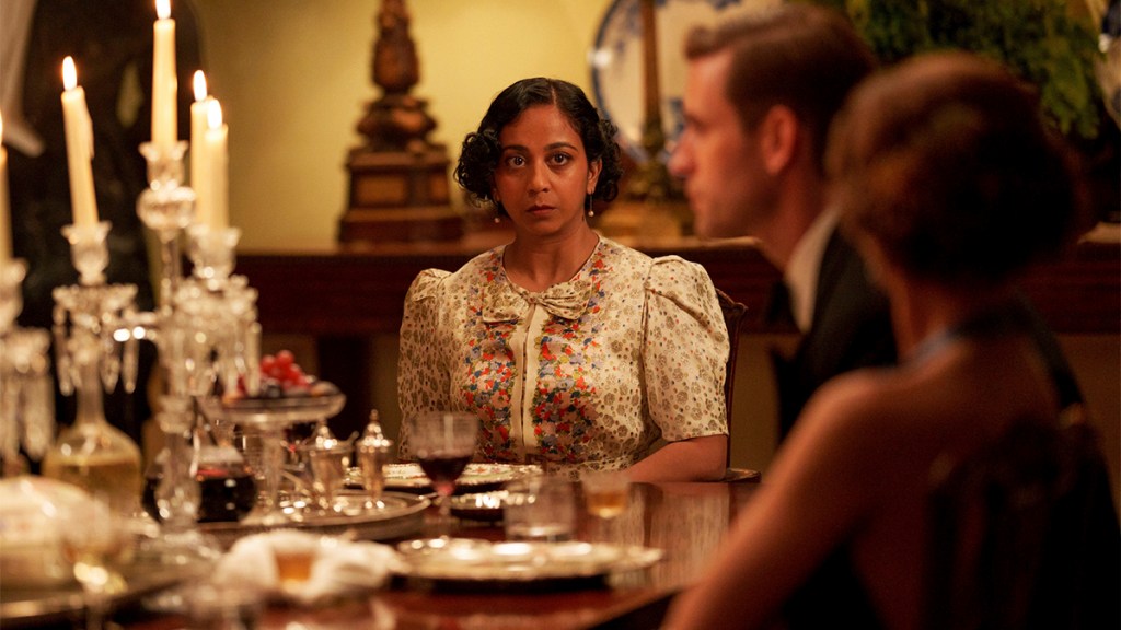 Anjana Vasan in a 1920s-style blouse sitting at the head of a dinner table