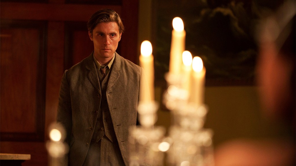 Jack Farthing in a 1920s-style suit standing by some candles