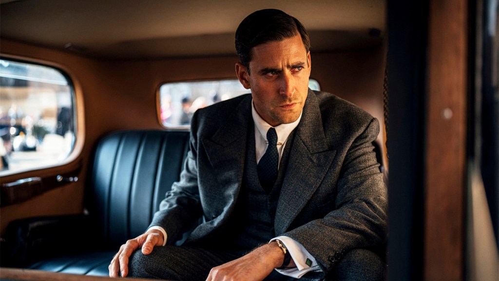 Oliver Jackson-Cohen in a suit in the back of a 1920s-style car