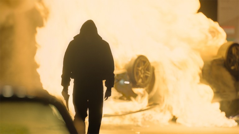 A silhouetted man walking away from a burning car in Gangs of London series 3