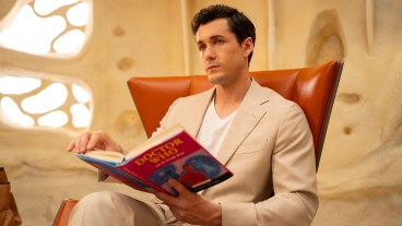 Jonah Hauer-King as Conrad Clark in a cream-coloured suit sitting in a chair and reading a Doctor Who book