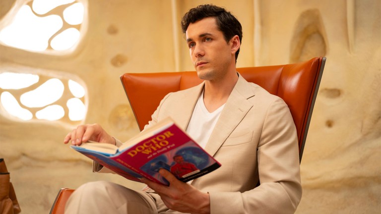 Jonah Hauer-King as Conrad Clark in a cream-coloured suit sitting in a chair and reading a Doctor Who book